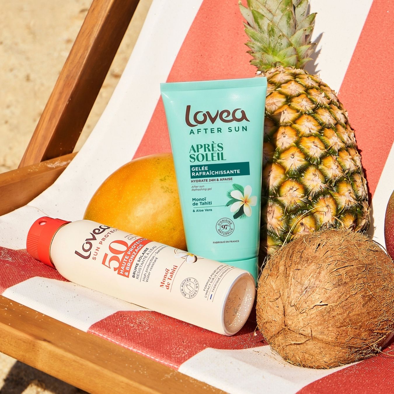 Lovea Duo Solaire Bronzage Doré – Active Protège & Prolonge au monoï de Tahiti, idéal pour des soins solaires haut de gamme et un bronzage doré éclatant, repose sur une chaise longue rayée de rouge et de blanc, à côté d’un ananas, d’une noix de coco et d’une mangue, sur du sable baigné de soleil.