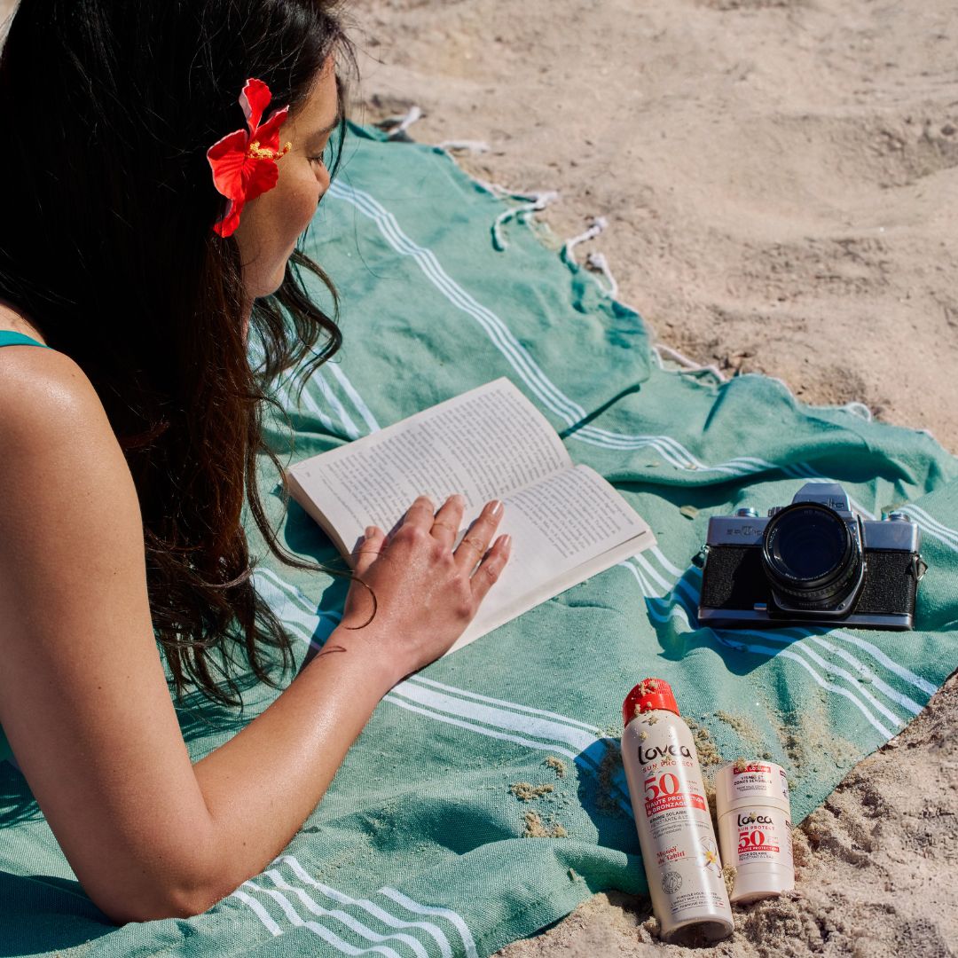 Une femme, une fleur rouge dans les cheveux, lit un livre allongée sur une serviette rayée verte à la plage. À côté d'elle se trouvent un appareil photo et deux flacons de crème solaire indice 50.